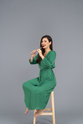 In voller länge einer lächelnden jungen dame im grünen kleid, die auf einem stuhl sitzt, während sie klarinette spielt