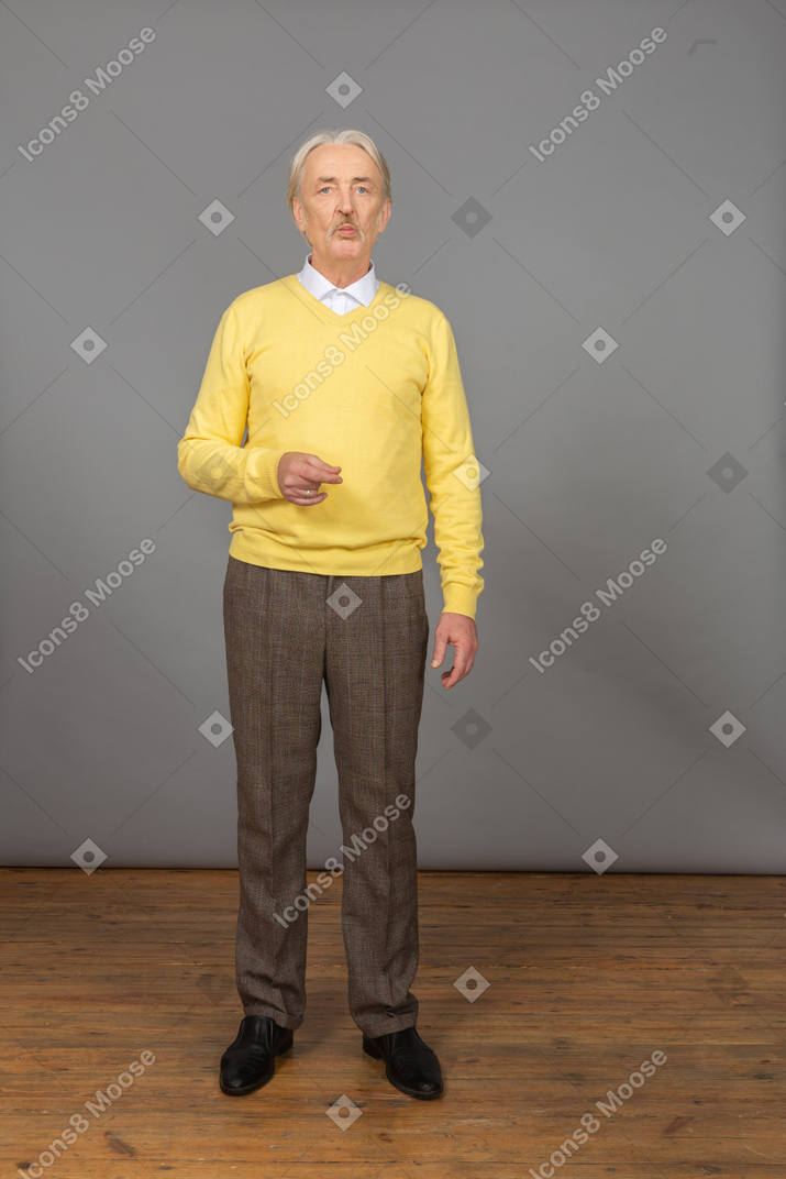 Vista frontal de um velho perplexo com um pulôver amarelo, levantando a mão e olhando para a câmera