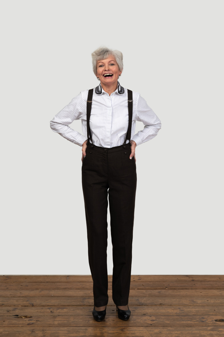 An old female standing on a wooden floor dressed in trousers with suspenders and laughing putting hands on hips