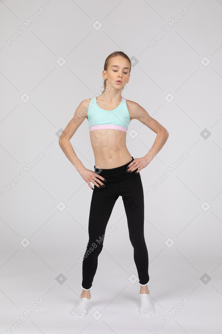 一个年轻的女孩，穿着运动服，将手放在臀部上，往下看的前视图