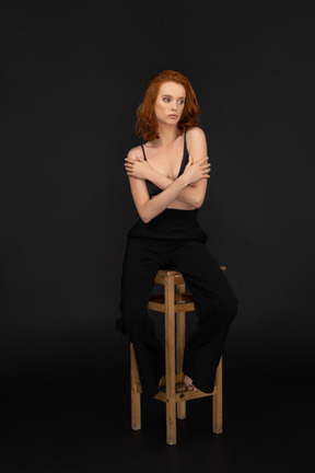 A frontal view of the sexy elegant woman, sitting on the wooden chair, hugging herself and looking to the right