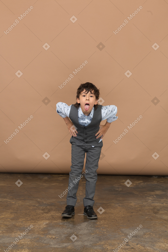 Вид спереди мальчика в костюме, стоящего с руками на бедрах и наклоняющегося вниз