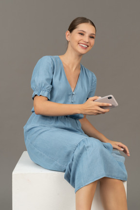 Vue de trois quarts d'une jeune femme assise sur un cube et souriant avec un smartphone à la main