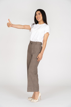 Vista de três quartos de uma jovem sorridente de calça e camiseta mostrando o polegar para cima
