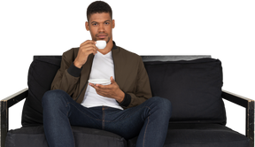 Front view of a young dreaming man sitting on a sofa while drinking coffee