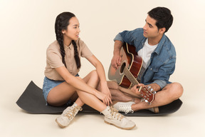 Young caucasian guy playing guitar sitting near asian woman on karimat