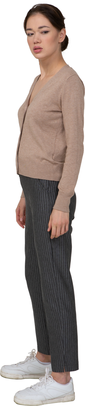 Вид в три четверти угрюмой молодой леди, стоящей неподвижно в пуловере и штанах и смотрящей в сторону