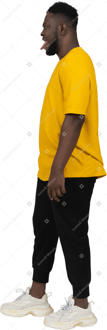 一个身穿黄色 t 恤的黑皮肤年轻男子站着不动并露出舌头的侧视图