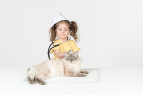 Criança menina com estetoscópio e usando chapéu médico curando um gato