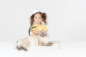 Little kid girl with stethoscope and wearing medical hat curing a cat