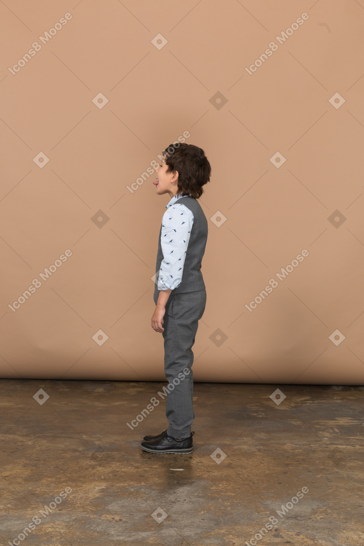 Вид сбоку симпатичного мальчика в сером костюме, показывающего язык
