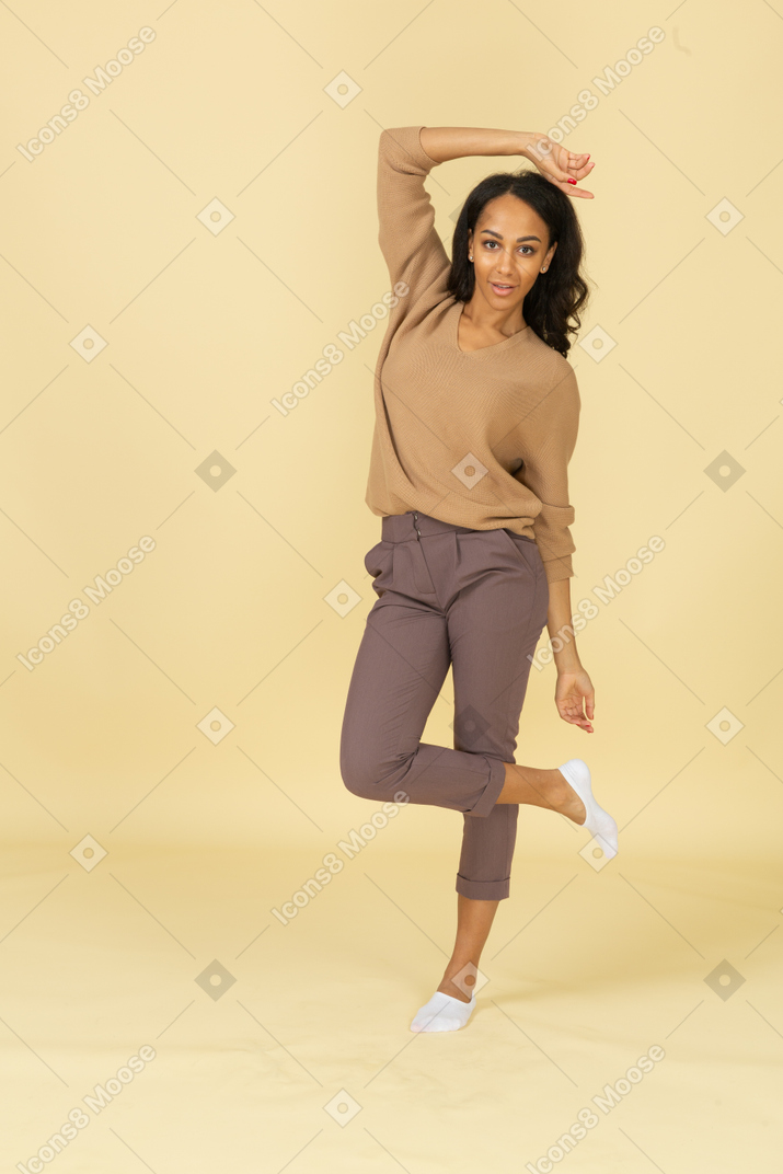 Vista frontal de una mujer joven de piel oscura tocando la cabeza mientras levanta la pierna