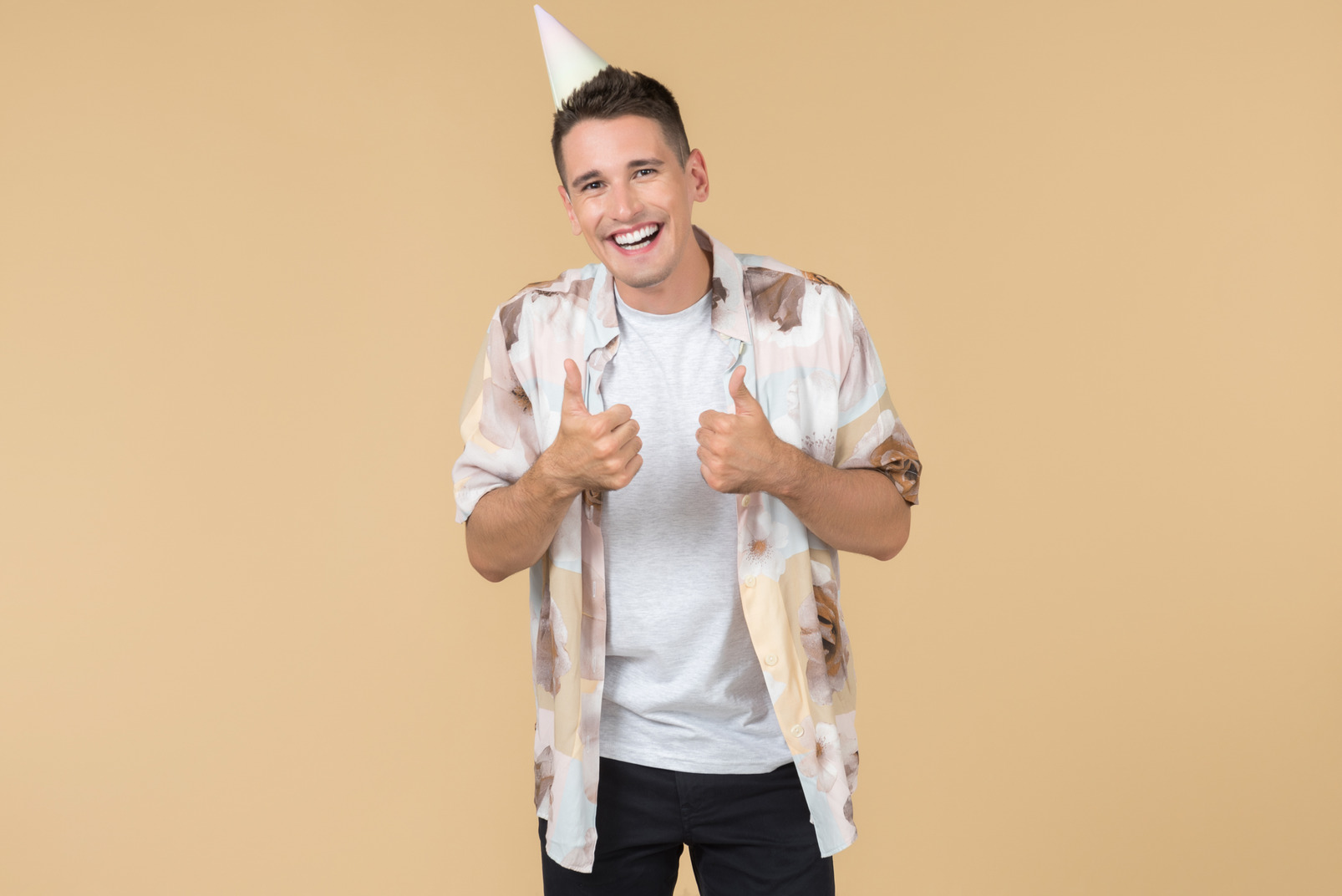 Laughing young party guy with birthday cone on the head showing thumb up