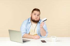 Junger übergewichtiger mann, der am tisch sitzt, telefon hält und scheckkarte betrachtet