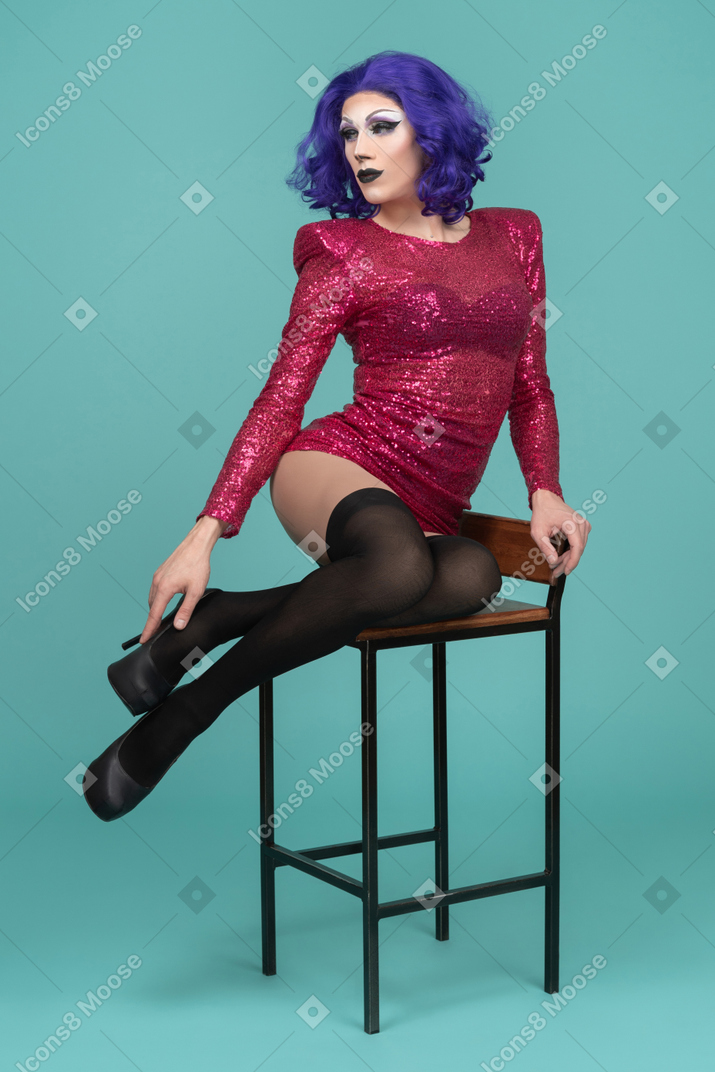 Drag queen sentada en un taburete con las piernas dobladas y tocando el zapato