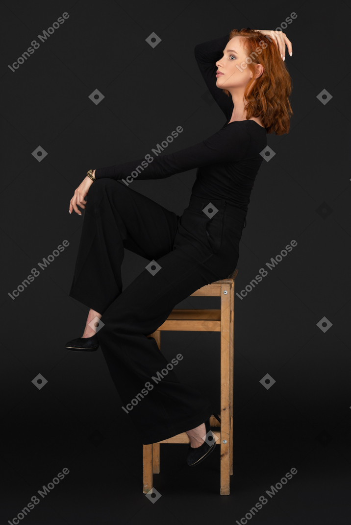 A side view of the young beautiful woman sitting on the tall woden chair and adjusting her hair