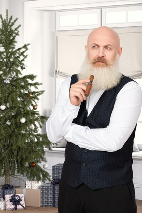 A bald man in a vest holding a cigar next to a christmas tree