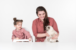 Mother and her little daughter, wearing red and pink clothes, sitting at the dinner table with their family cat