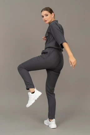 Three-quarter back view of a young woman in a jumpsuit raising hand and leg