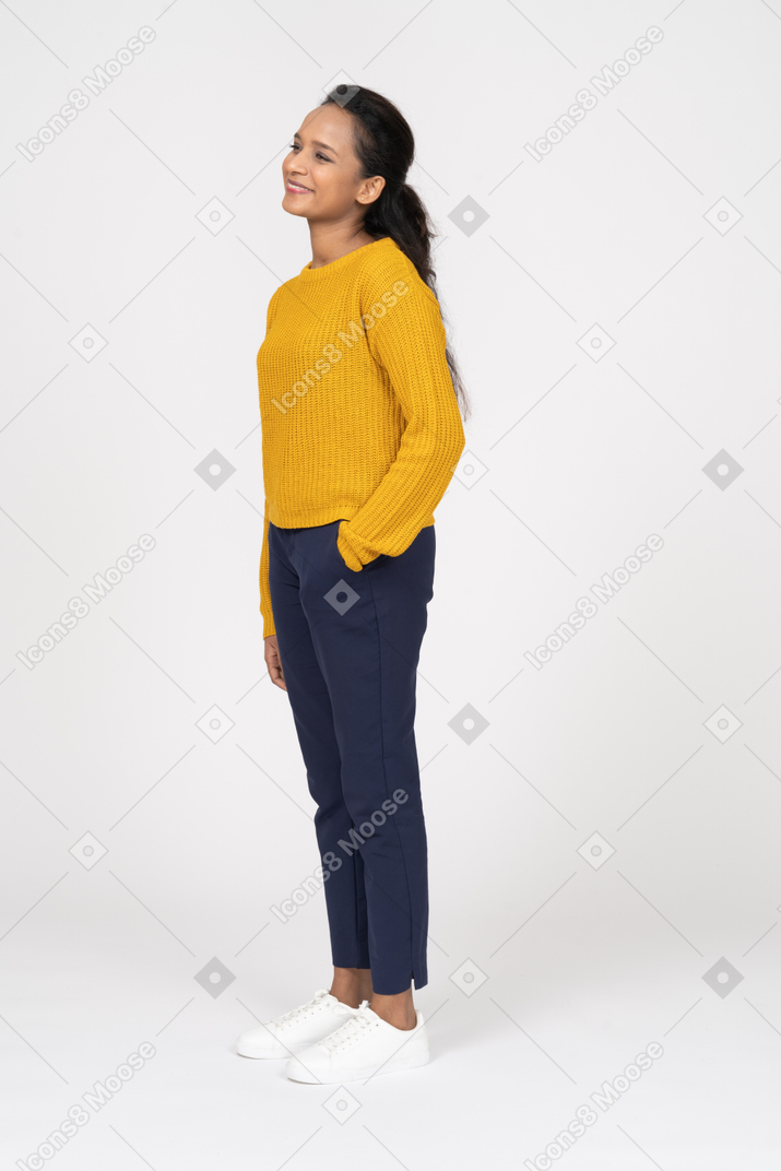 Vista lateral de una niña feliz en ropa casual posando con la mano en el bolsillo