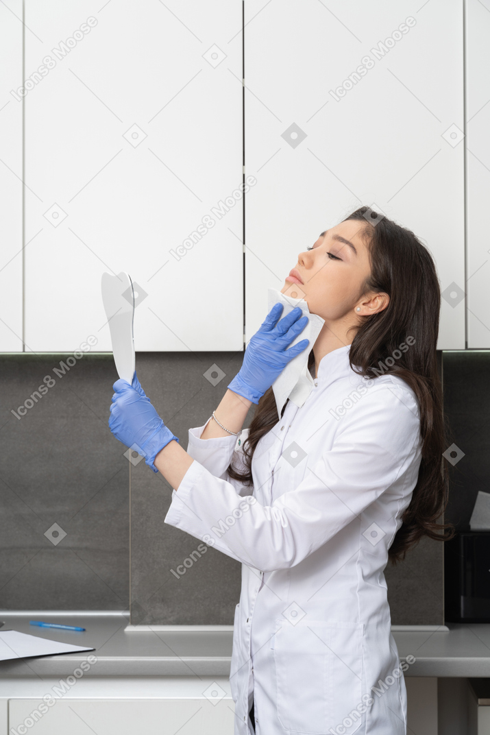 Vista lateral de una doctora sosteniendo un espejo y secándose la cara