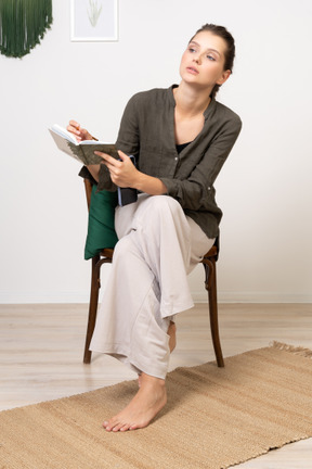Вид спереди вдумчивой молодой женщины в домашней одежде, сидящей на стуле с карандашом и блокнотом