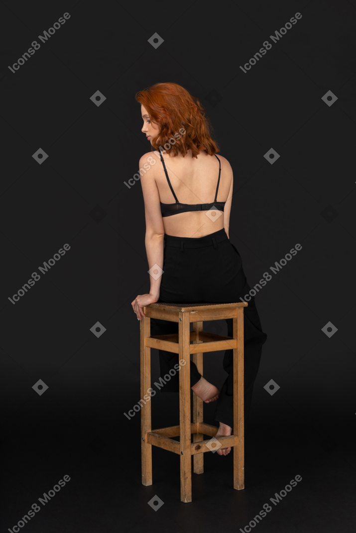 A back side of the beautiful woman dressed in black pants and bra, sitting on the wooden chair and looking to the left