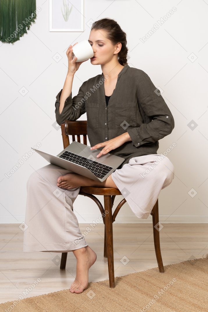 Vista frontal de una mujer joven vistiendo ropa de casa sentado en una silla con una computadora portátil y tomando café