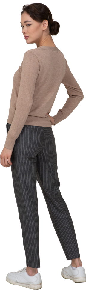 Vista posterior de tres cuartos de una mujer astuta en suéter y pantalones poniendo la mano en la cadera