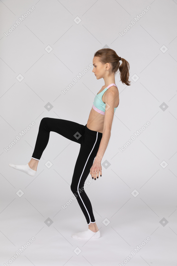 Side view of a teen girl in sportswear outspreading hands and raising leg