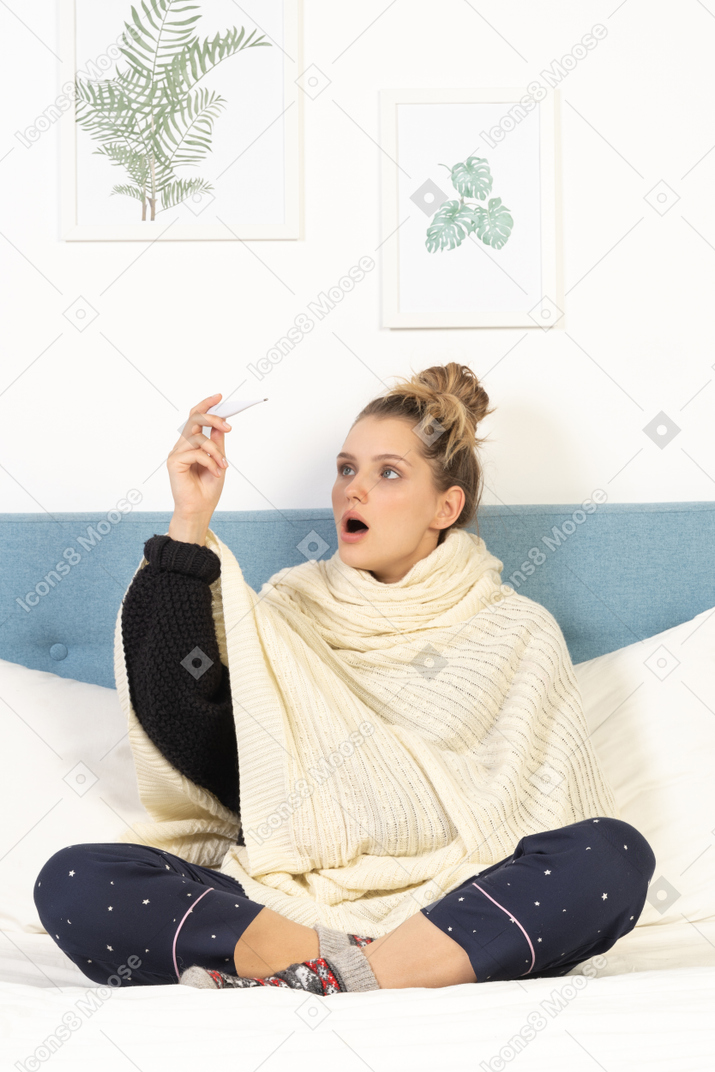 一位震惊的年轻女士裹着白色毯子坐在床上，带着温度计的前视图