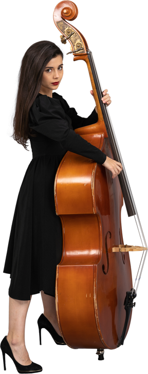 Vista laterale di un serio giovane musicista femminile in abito nero che suona il suo contrabbasso