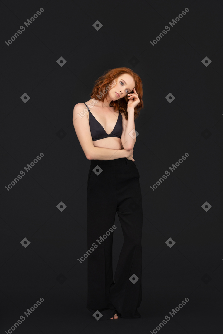 A frontal view of the cute red haired girl posing on the black background touching her hair and looking to the right