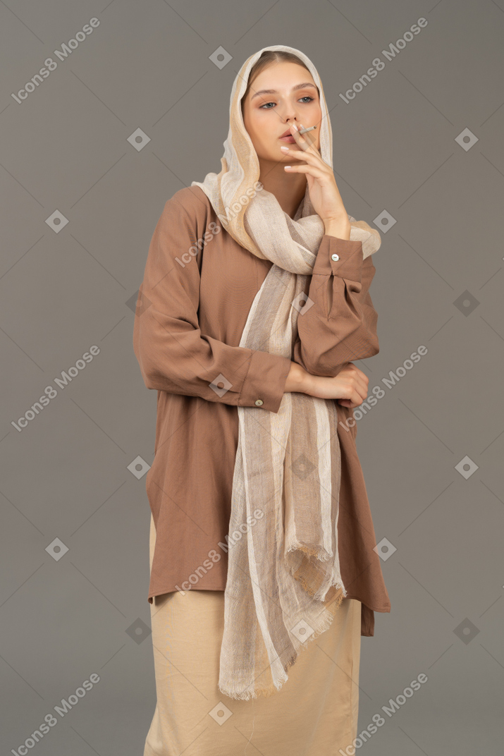 Woman in headscarf and beige clothes smoking a cigarette and looking aside