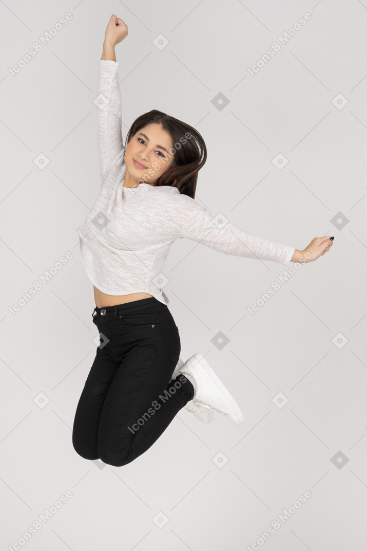 Three-quarter view of a smiling jumping young indian female in casual clothes raising hand
