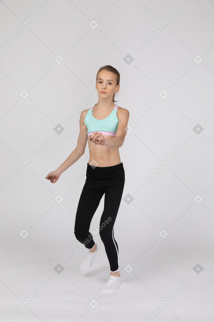 Three-quarter view of a teen girl in sportswear looking at camera while stepping forward and raising hand