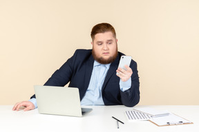 Pensieroso giovane uomo in sovrappeso seduto alla scrivania e in possesso di telefono