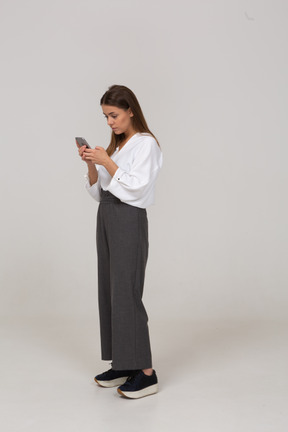 Vista de três quartos de uma jovem com roupas de escritório, verificando o feed por telefone