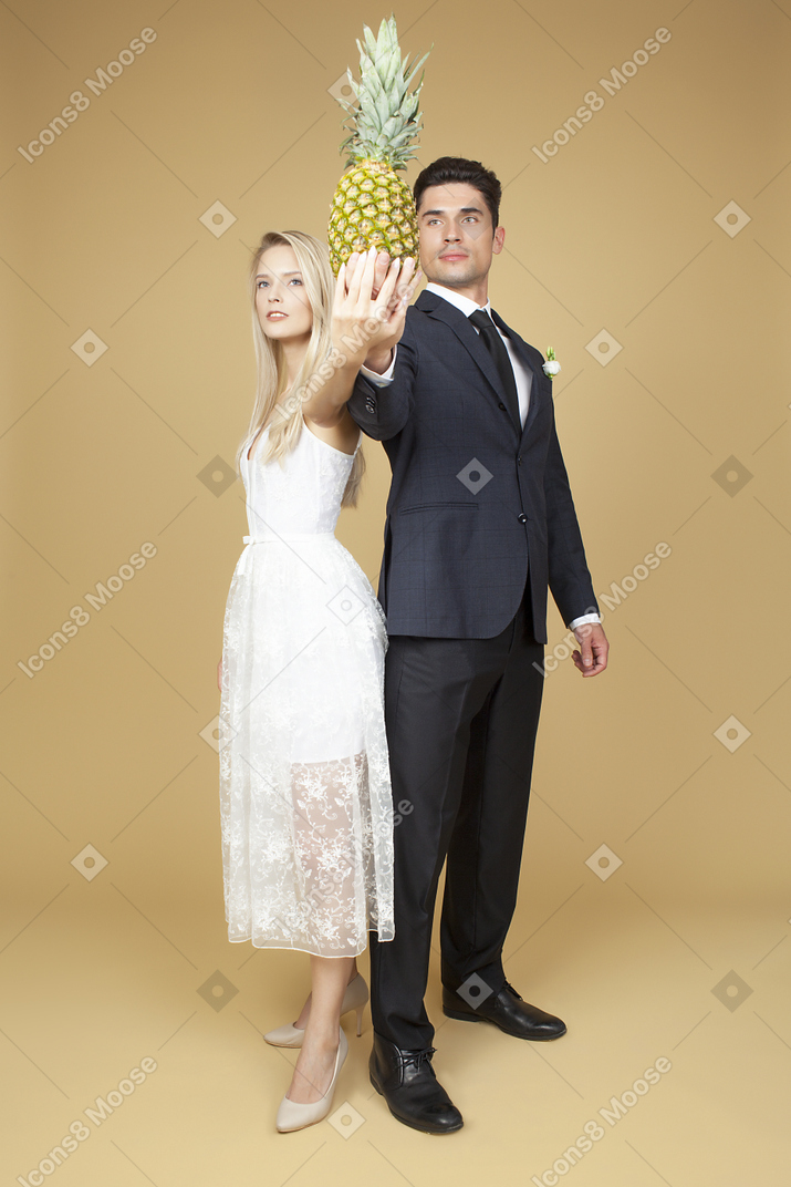 Groom and bride standing shoulder to shoulder and holding a pineapple up