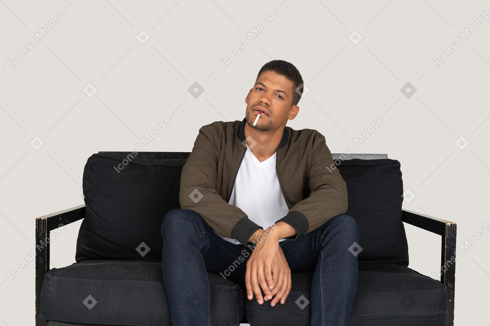 Front view of young man sitting on a sofa and holding cigarette in mouth