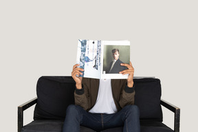 Front view of young man sitting on a sofa and holding a magazine
