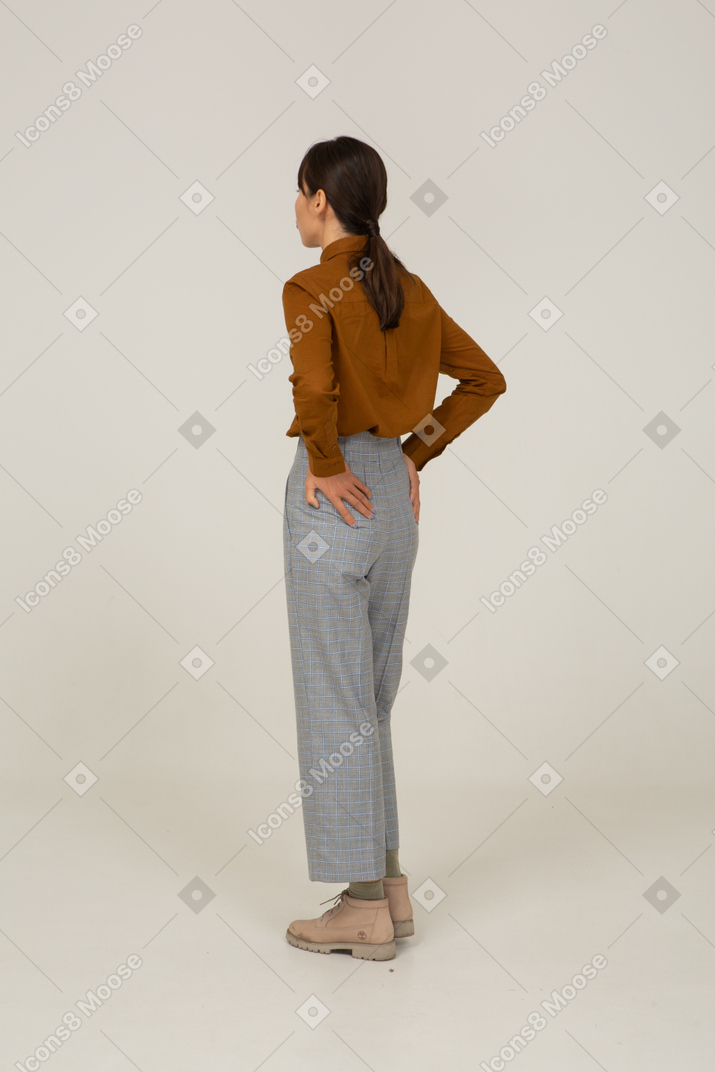 Vista posterior de tres cuartos de una joven mujer asiática haciendo pucheros en calzones y blusa poniendo las manos en las caderas