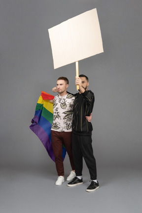 Vista de tres cuartos de dos hombres jóvenes sosteniendo una cartelera y una bandera lgbt