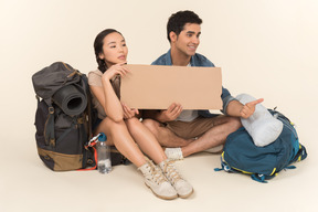 Young interracial couple of hitchhikers holding paper card and showing hitchhiking gestures