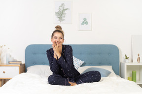 Front view of a pleased young woman in pajamas staying in bed and holding hands together