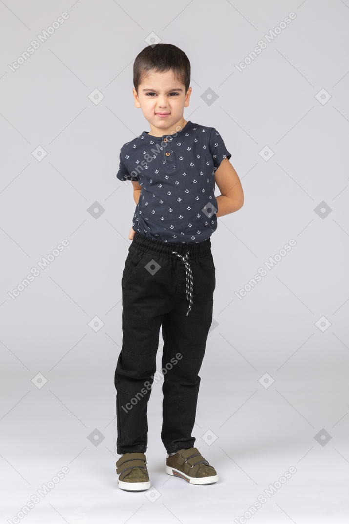 Vue de face d'un garçon en colère dans des vêtements décontractés posant avec les mains derrière le dos et regardant la caméra