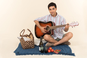 Junger kaukasischer kerl, der nahe picknickkorb auf der decke sitzt und gitarre spielt