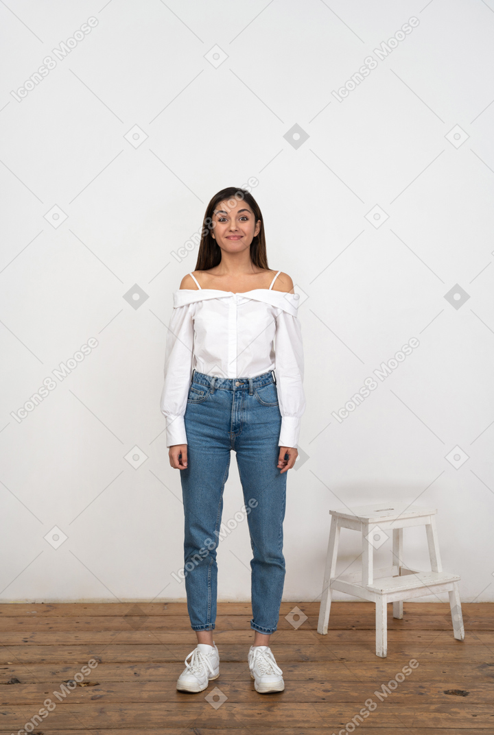 A young woman standing next to a white chair in front of a white wall