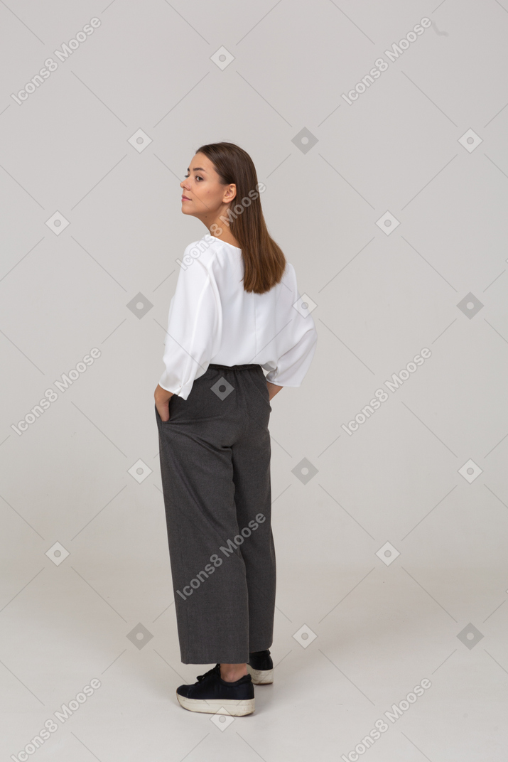 Three-quarter back view of a young lady in office clothing putting hands in pockets & looking aside