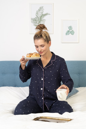 Vista frontal de una joven en pijama sosteniendo una taza de café y algunos pasteles sentado en la cama